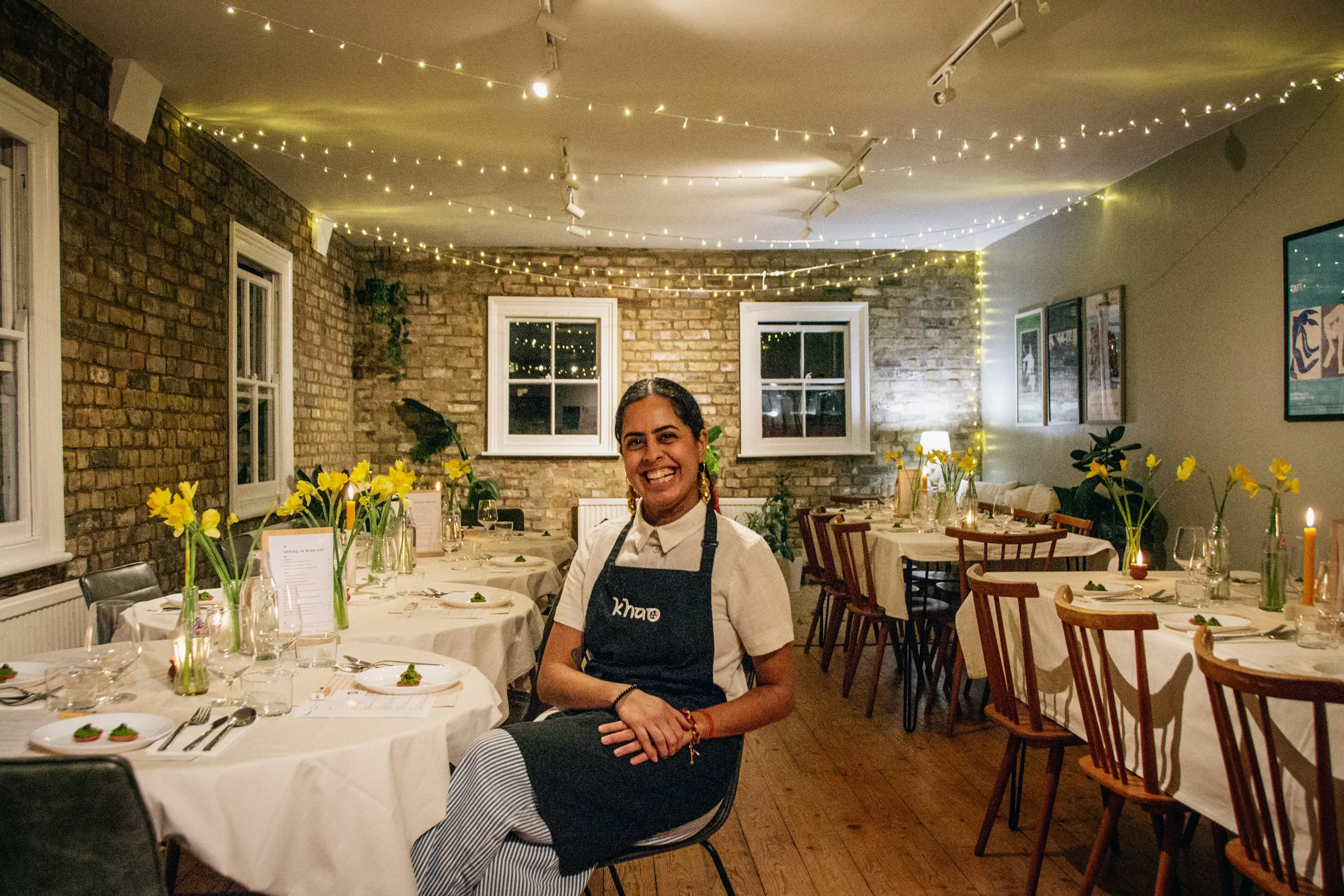 Smiling woman in a restaurant wearing a dark apron, sitting in front of tables decorated with yellow flowers and string lights.