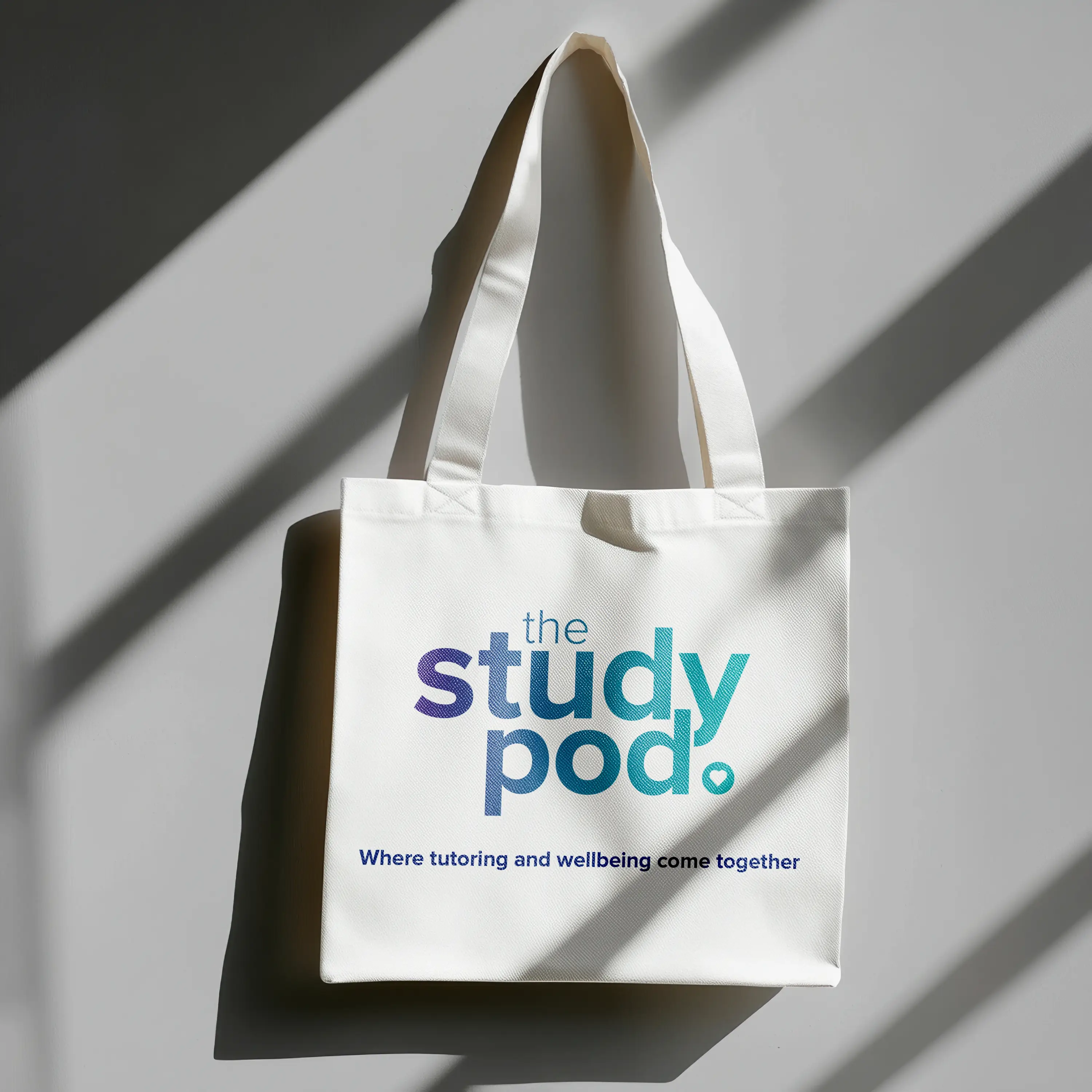 White tote bag with handles featuring the text 'the study pod. Where tutoring and wellbeing come together' on a gray surface with diagonal shadows.